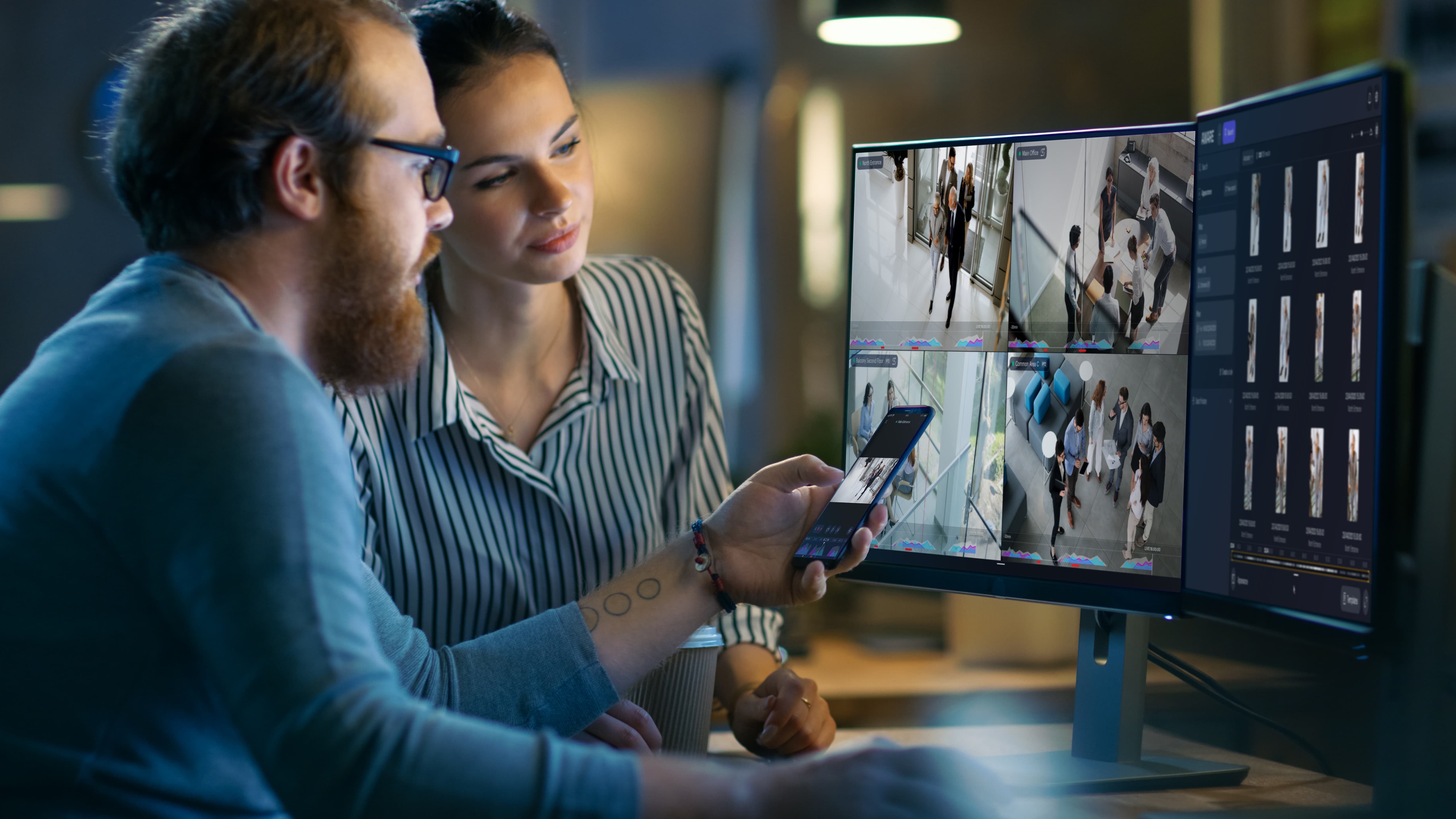 Two people monitoring multiple surveillance feeds on a computer and phone in a dim workspace.